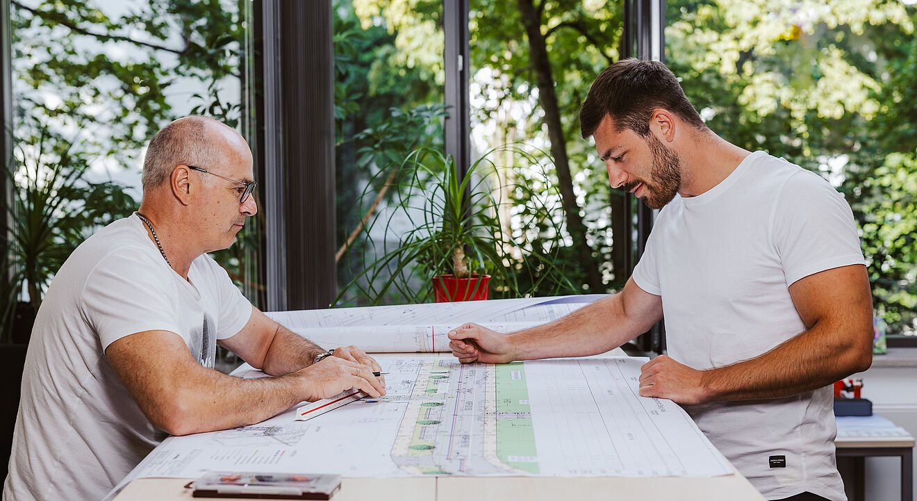 Zwei Personen sitzen an einem großen Tisch und betrachten technische Baupläne. Auf dem Tisch liegen mehrere großformatige Pläne mit detaillierten Zeichnungen und Markierungen. Im Hintergrund sind große Fenster mit Blick ins Grüne sowie eine rote Pflanze auf der Fensterbank sichtbar. Die Szene spielt in einem hellen Büro oder Besprechungsraum. Verlinkung zur Aufmassportal Seite.