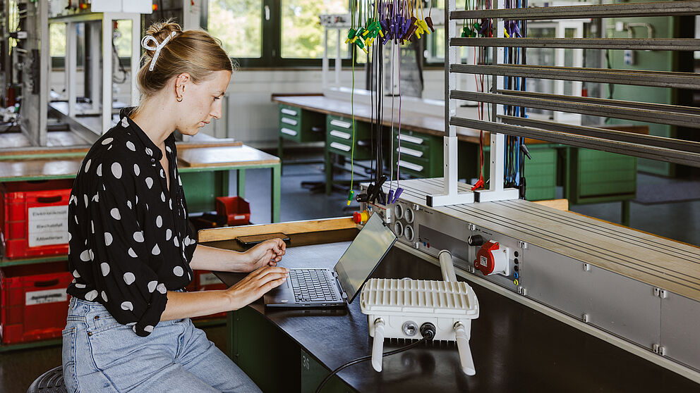 Person arbeitet in einem modernen Elektrotechnik-Labor an einem Laptop. Auf dem Arbeitstisch steht ein weißes Prüfgerät mit Kühlrippen und Kabelanschlüssen. Im Hintergrund sind grüne Werkbänke mit Schubladen, große Fenster für Tageslicht sowie technische Vorrichtungen mit bunten Kabeln sichtbar. Die Umgebung ist auf Montage- und Testarbeiten in der Energie- und Netztechnik ausgerichtet.