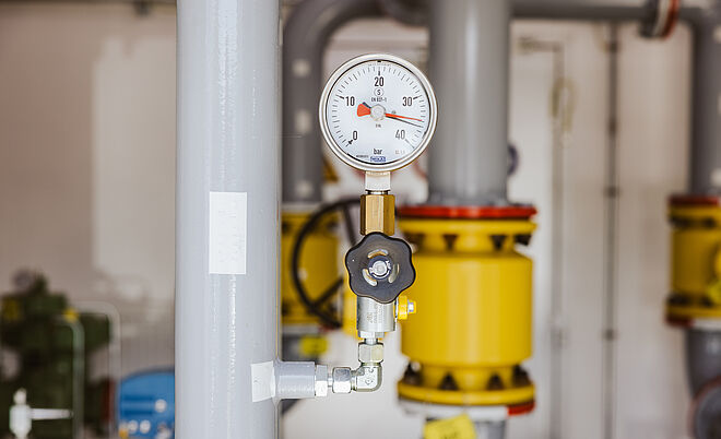 Nahaufnahme eines Manometers an einer grauen Rohrleitung in einem Technikraum. Das Druckmessgerät zeigt einen Bereich von 0 bis 30 bar. Im Hintergrund sind weitere graue Rohre mit gelben Armaturen und metallischen Verbindungen sichtbar. Verlinkung zur Netzanschluss Gas Seite.
