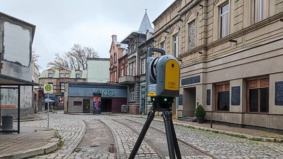 Vermessung einer historischen Straßenkulisse mit Kopfsteinpflaster und Straßenbahnschienen. Im Vordergrund steht ein modernes Laserscanning-Gerät auf einem Stativ, ausgerichtet auf die Umgebung. Im Hintergrund sind alte Gebäude mit aufwendigen Fassaden, darunter ein Kino mit blauer Beschriftung, zu sehen. Links befindet sich ein Bushaltestellenschild. Die Szene zeigt eine präzise Vermessung für digitale Erfassung und 3D-Modellierung.