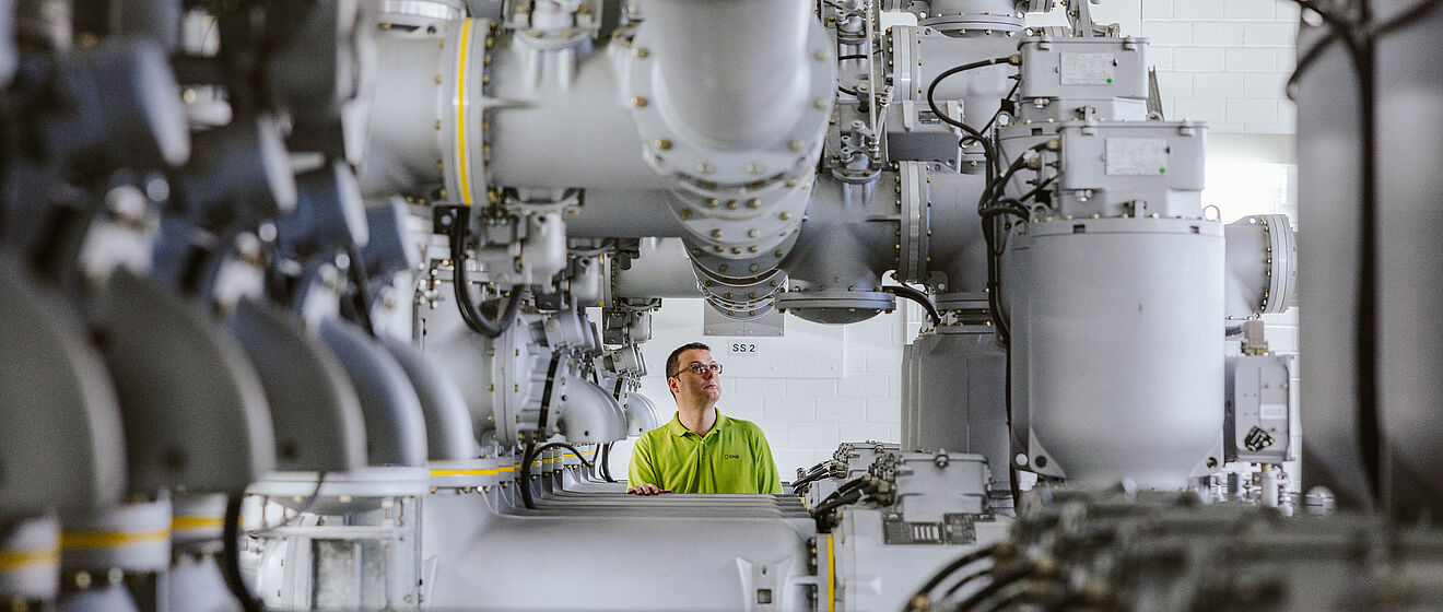 Innenansicht einer gasisolierten Schaltanlage (GIS) in einem Umspannwerk: Mehrere große, silberfarbene Hochspannungsrohre und Schaltkomponenten verlaufen parallel und sind mit Ventilen, Isolatoren und Verbindungselementen ausgestattet. Im Hintergrund steht eine Person in Warnschutzkleidung zwischen den Anlagen. Die Szene zeigt moderne Hochspannungstechnik für Stromversorgung und Netzbetrieb. Verlinkung zur Auswahl Einspeisung Seite.