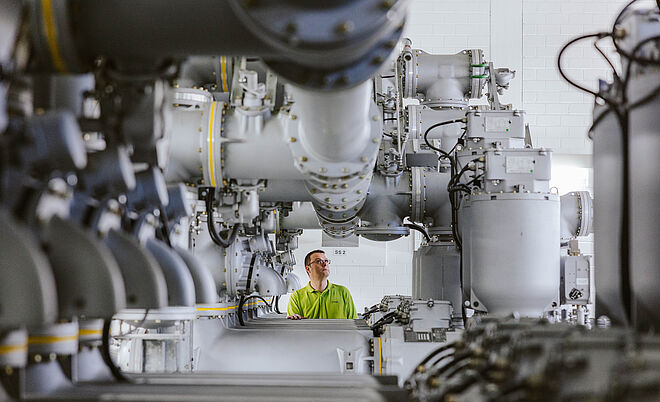 Innenansicht einer gasisolierten Schaltanlage (GIS) in einem Umspannwerk: Mehrere große, silberfarbene Hochspannungsrohre und Schaltkomponenten verlaufen parallel und sind mit Ventilen, Isolatoren und Verbindungselementen ausgestattet. Im Hintergrund steht eine Person in Warnschutzkleidung zwischen den Anlagen. Die Szene zeigt moderne Hochspannungstechnik für Stromversorgung und Netzbetrieb. Verlinkung zur Auswahl Einspeisung Seite.