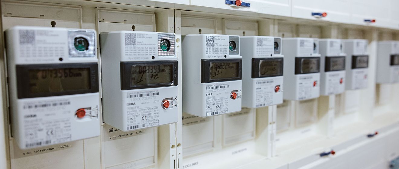 Mehrere digitale Stromzähler sind in einer weißen Schalttafel nebeneinander installiert. Jeder Zähler verfügt über ein Display mit Messwerten, einen roten Bedientaster und einen QR-Code in der oberen Ecke. Die Geräte sind sauber angeordnet und zeigen eine moderne Infrastruktur für Energie- und Verbrauchsmessung. Die Szene verdeutlicht den Einsatz von Smart-Meter-Technologie für präzise Stromabrechnung und Netzüberwachung.