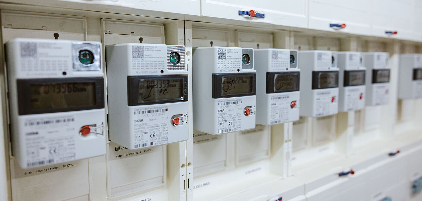 Mehrere digitale Stromzähler sind in einer weißen Schalttafel nebeneinander installiert. Jeder Zähler verfügt über ein Display mit Messwerten, einen roten Bedientaster und einen QR-Code in der oberen Ecke. Die Geräte sind sauber angeordnet und zeigen eine moderne Infrastruktur für Energie- und Verbrauchsmessung. Die Szene verdeutlicht den Einsatz von Smart-Meter-Technologie für präzise Stromabrechnung und Netzüberwachung.