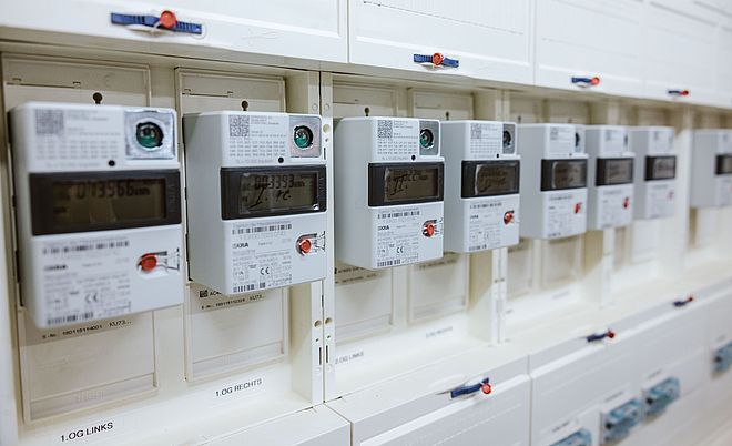 Mehrere digitale Stromzähler sind in einer weißen Schalttafel nebeneinander installiert. Jeder Zähler verfügt über ein Display mit Messwerten, einen roten Bedientaster und einen QR-Code in der oberen Ecke. Die Geräte sind sauber angeordnet und zeigen eine moderne Infrastruktur für Energie- und Verbrauchsmessung. Die Szene verdeutlicht den Einsatz von Smart-Meter-Technologie für präzise Stromabrechnung und Netzüberwachung. Verlinkung zur Seite Zähleranschlusssäulen.