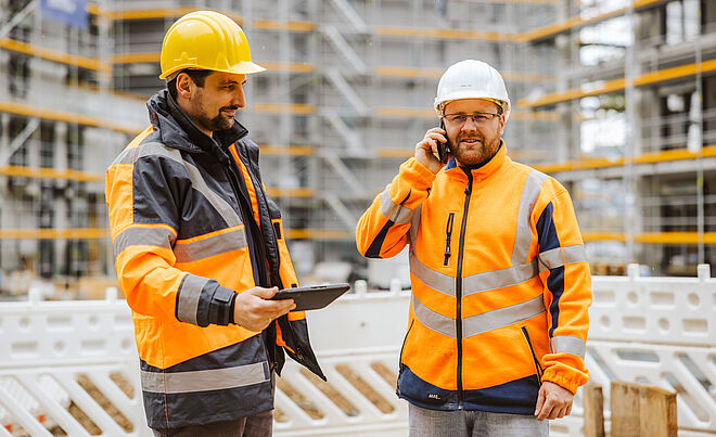 Zwei Personen in Sicherheitskleidung stehen auf einer Baustelle vor einem Gebäude mit Gerüsten. Beide tragen orange Warnschutzjacken mit reflektierenden Streifen und Schutzhelme. Eine Person hält ein Tablet in der Hand, die andere telefoniert. Im Hintergrund sind Baugerüste und Absperrungen sichtbar, was auf aktive Bauarbeiten hinweist. Die Szene zeigt Koordination und Kommunikation bei einem Infrastrukturprojekt. Verlinkung zur Bau- und Veranstaltungsstrom Seite.