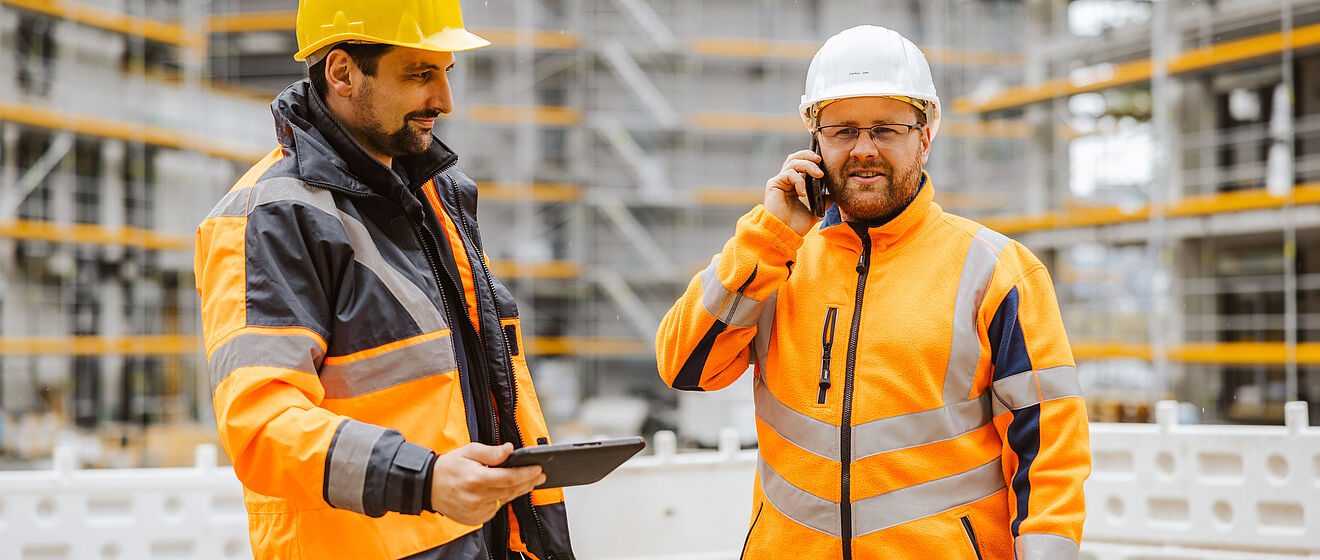 Zwei Personen in Sicherheitskleidung stehen auf einer Baustelle vor einem Gebäude mit Gerüsten. Beide tragen orange Warnschutzjacken mit reflektierenden Streifen und Schutzhelme. Eine Person hält ein Tablet in der Hand, die andere telefoniert. Im Hintergrund sind Baugerüste und Absperrungen sichtbar, was auf aktive Bauarbeiten hinweist. Die Szene zeigt Koordination und Kommunikation bei einem Infrastrukturprojekt. Verlinkung zur Bau- und Veranstaltungsstrom Seite.