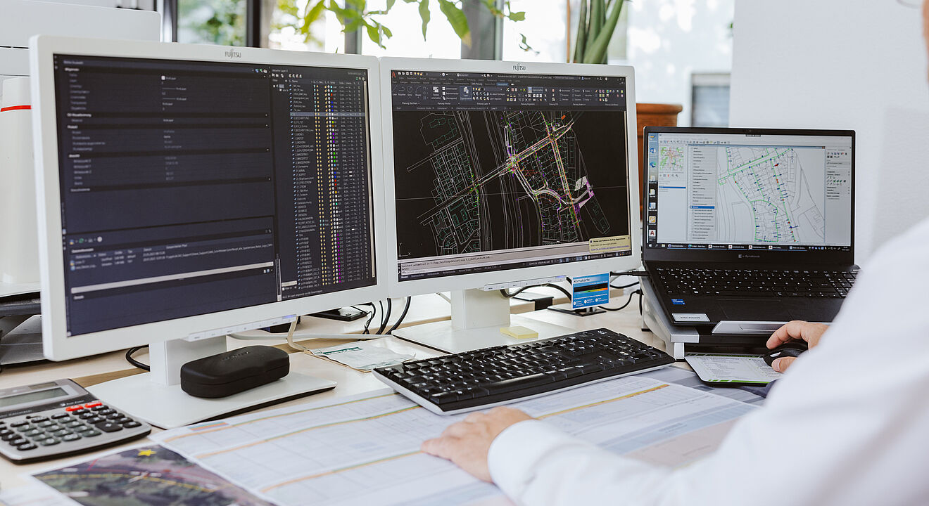 Arbeitsplatz mit drei Bildschirmen, auf denen technische Planungssoftware geöffnet ist. Die beiden großen Monitore zeigen detaillierte Leitungs- und Netzpläne in CAD-Ansicht mit farbigen Linien und Datenlisten. Der Laptop rechts zeigt ebenfalls eine Netzskizze. Auf dem Tisch liegen großformatige technische Pläne, daneben eine Tastatur, Maus und ein Taschenrechner. Im Hintergrund sind Fenster mit Tageslicht und grüne Pflanzen sichtbar. Die Szene verdeutlicht digitale Planung und Dokumentation für Infrastrukturprojekte. Verlinkung zur Veröffentlichungen Allgemein Seite.