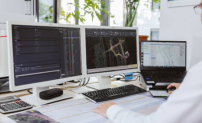 Arbeitsplatz mit drei Bildschirmen, auf denen technische Planungssoftware geöffnet ist. Die beiden großen Monitore zeigen detaillierte Leitungs- und Netzpläne in CAD-Ansicht mit farbigen Linien und Datenlisten. Der Laptop rechts zeigt ebenfalls eine Netzskizze. Auf dem Tisch liegen großformatige technische Pläne, daneben eine Tastatur, Maus und ein Taschenrechner. Im Hintergrund sind Fenster mit Tageslicht und grüne Pflanzen sichtbar. Die Szene verdeutlicht digitale Planung und Dokumentation für Infrastrukturprojekte. Verlinkung zur Veröffentlichungen Allgemein Seite.