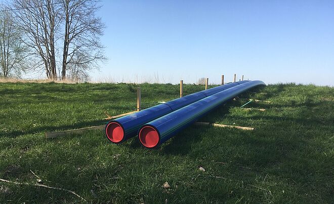Zwei lange blaue Rohre mit roten Endkappen liegen parallel auf einer grünen Wiese. Die Rohre sind auf Holzstützen erhöht abgelegt. Im Hintergrund stehen einige kahle Bäume, und der Himmel ist klar und blau. Die Szene wirkt wie eine Baustellenvorbereitung für Rohrleitungsarbeiten im Freien. Verlinkung zur Erschliessungsgebiete Seite.