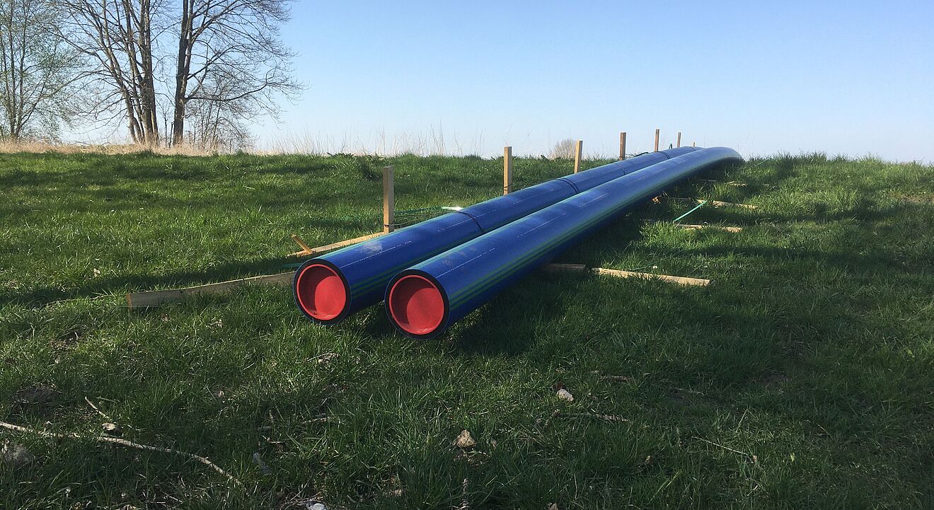 Zwei lange blaue Rohre mit roten Endkappen liegen parallel auf einer grünen Wiese. Die Rohre sind auf Holzstützen erhöht abgelegt. Im Hintergrund stehen einige kahle Bäume, und der Himmel ist klar und blau. Die Szene wirkt wie eine Baustellenvorbereitung für Rohrleitungsarbeiten im Freien. Verlinkung zur Erschliessungsgebiete Seite.