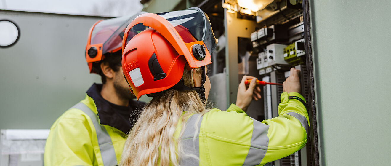 Zwei Personen in Warnschutzkleidung und roten Schutzhelmen arbeiten an einer geöffneten Schaltanlage in einem Umspannwerk. Eine Person hält einen Schraubendreher und führt eine Einstellung oder Wartung an elektrischen Komponenten durch. Im Inneren der Anlage sind Schalter, Kabel und Messgeräte sichtbar, darüber eine Lampe zur Beleuchtung. Die Szene zeigt sichere Arbeiten an Hochspannungsanlagen im Bereich Stromversorgung und Netztechnik. Verlinkung zur Netzqualitätsmessung Seite.