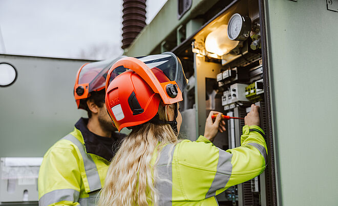 Zwei Personen in Warnschutzkleidung und roten Schutzhelmen arbeiten an einer geöffneten Schaltanlage in einem Umspannwerk. Eine Person hält einen Schraubendreher und führt eine Einstellung oder Wartung an elektrischen Komponenten durch. Im Inneren der Anlage sind Schalter, Kabel und Messgeräte sichtbar, darüber eine Lampe zur Beleuchtung. Die Szene zeigt sichere Arbeiten an Hochspannungsanlagen im Bereich Stromversorgung und Netztechnik. Verlinkung zur Netzqualitätsmessung Seite.