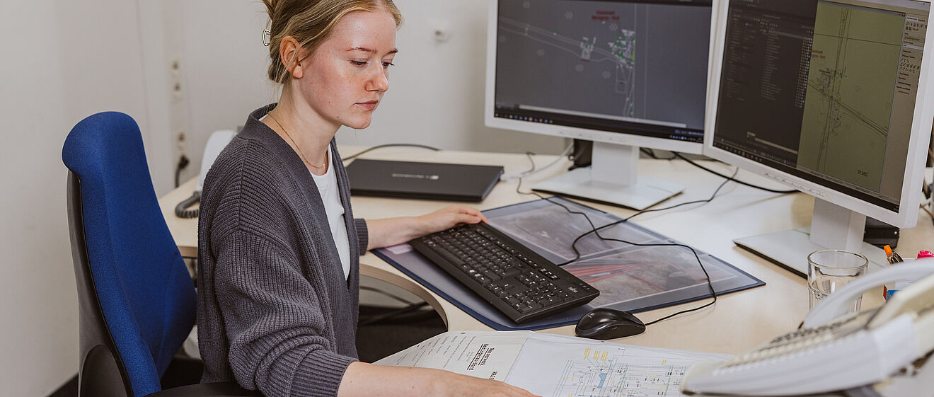 Eine Person sitzt an einem Schreibtisch und arbeitet an einem Computer mit zwei großen Monitoren. Auf den Bildschirmen sind technische Zeichnungen und Pläne zu sehen. Vor der Person liegt ein großformatiger Papierplan ausgebreitet. Auf dem Schreibtisch befinden sich außerdem eine Tastatur, eine Maus, ein Telefon und ein Glas Wasser. Im Hintergrund steht eine grüne Pflanze am Fenster, durch das Tageslicht einfällt. Verlinkung zur Leitungsplanauskunft Seite.
