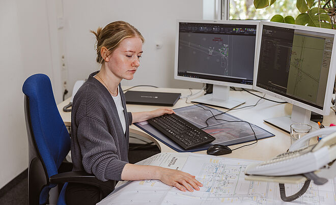 Eine Person sitzt an einem Schreibtisch und arbeitet an einem Computer mit zwei großen Monitoren. Auf den Bildschirmen sind technische Zeichnungen und Pläne zu sehen. Vor der Person liegt ein großformatiger Papierplan ausgebreitet. Auf dem Schreibtisch befinden sich außerdem eine Tastatur, eine Maus, ein Telefon und ein Glas Wasser. Im Hintergrund steht eine grüne Pflanze am Fenster, durch das Tageslicht einfällt. Verlinkung zur Leitungsplanauskunft Seite.