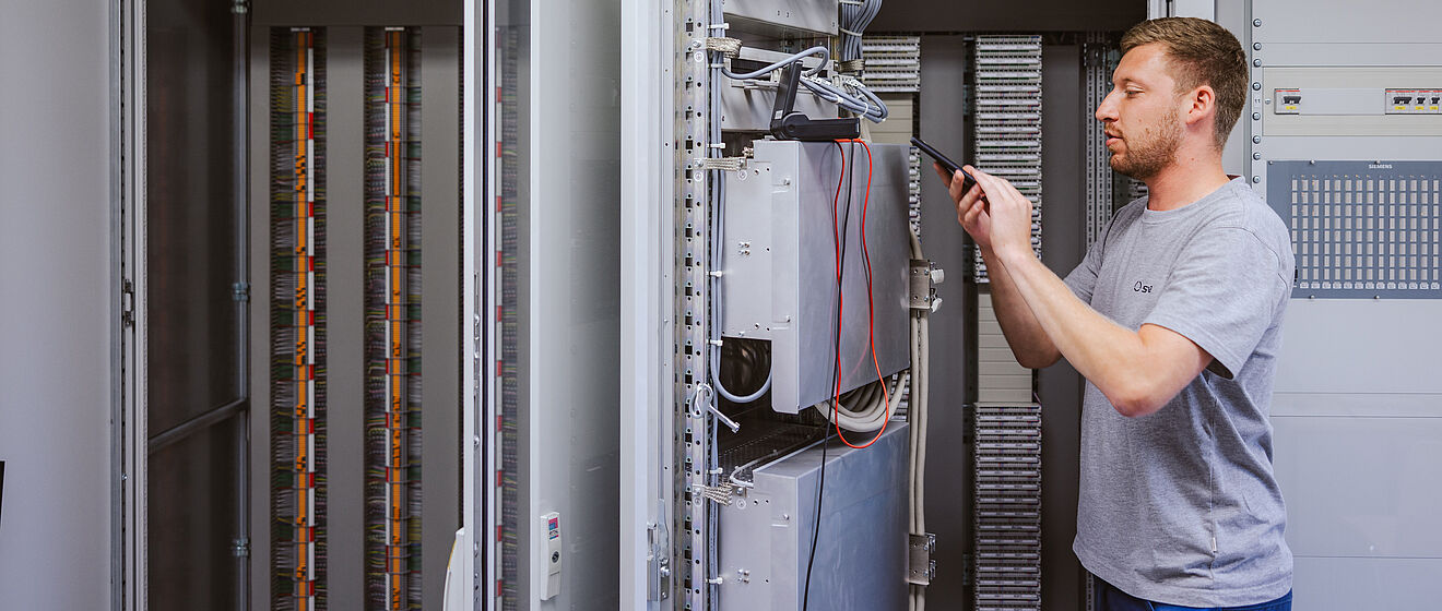 Techniker arbeitet an einem geöffneten Schaltschrank in einem Technikraum: Der Schaltschrank enthält komplexe elektrische Komponenten, Kabel und Steuergeräte. Der Techniker hält ein Werkzeug und führt eine Einstellung oder Wartung durch. Im Hintergrund sind weitere geöffnete Schaltschränke mit Kabelreihen sichtbar. An der Wand befindet sich ein Schild mit der Aufschrift ‚Siemens FW-Geräteschrank‘. Die Szene zeigt Arbeiten an Steuer- und Netztechnik für Energieversorgung. Verlinkung zur Schutzprüfung Seite.