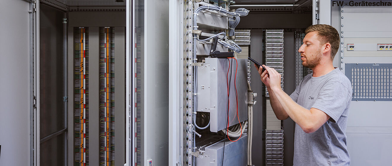 Techniker arbeitet an einem geöffneten Schaltschrank in einem Technikraum: Der Schaltschrank enthält komplexe elektrische Komponenten, Kabel und Steuergeräte. Der Techniker hält ein Werkzeug und führt eine Einstellung oder Wartung durch. Im Hintergrund sind weitere geöffnete Schaltschränke mit Kabelreihen sichtbar. An der Wand befindet sich ein Schild mit der Aufschrift ‚Siemens FW-Geräteschrank‘. Die Szene zeigt Arbeiten an Steuer- und Netztechnik für Energieversorgung. Verlinkung zur Schutzprüfung Seite.