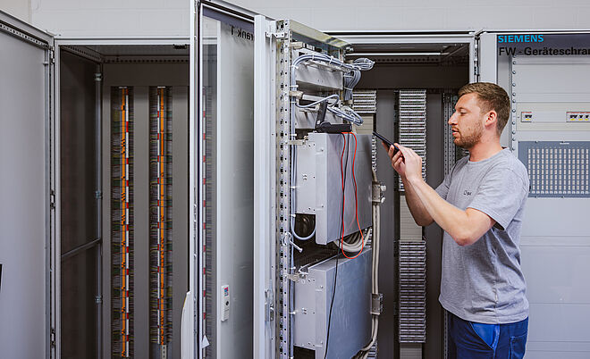 Techniker arbeitet an einem geöffneten Schaltschrank in einem Technikraum: Der Schaltschrank enthält komplexe elektrische Komponenten, Kabel und Steuergeräte. Der Techniker hält ein Werkzeug und führt eine Einstellung oder Wartung durch. Im Hintergrund sind weitere geöffnete Schaltschränke mit Kabelreihen sichtbar. An der Wand befindet sich ein Schild mit der Aufschrift ‚Siemens FW-Geräteschrank‘. Die Szene zeigt Arbeiten an Steuer- und Netztechnik für Energieversorgung. Verlinkung zur Schutzprüfung Seite.