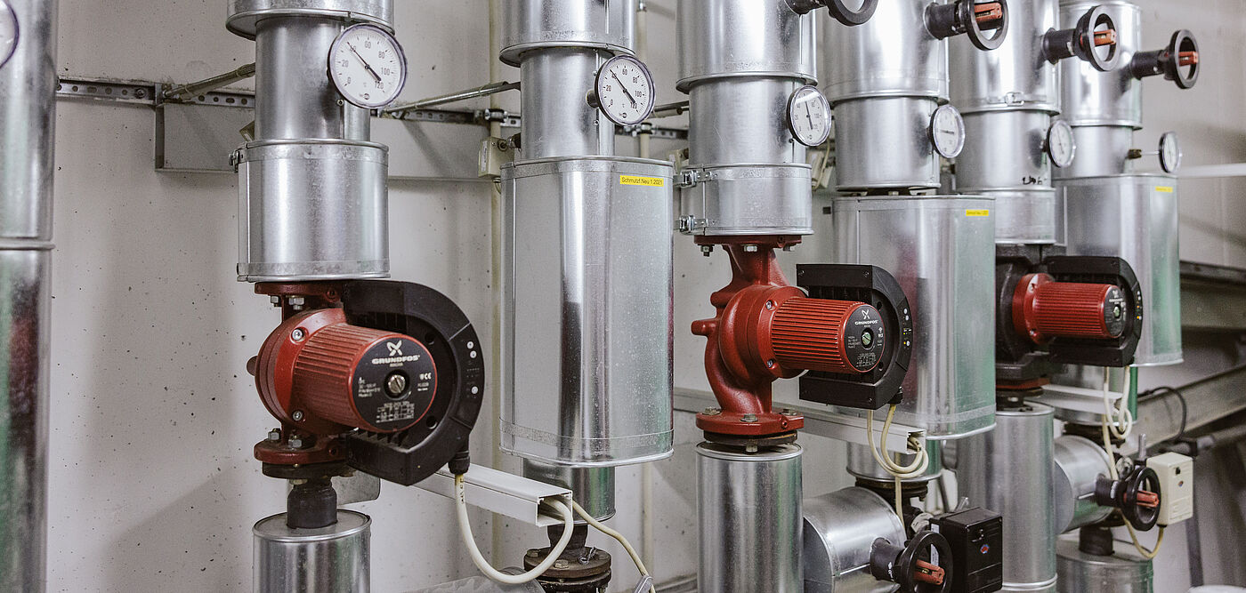 Mehrere silberne Rohrleitungen mit roten Umwälzpumpen und integrierten Druck- sowie Temperaturanzeigen in einem Technikraum. Die Anlage besteht aus isolierten Metallrohren, Manometern und elektrischen Steuerungen, typisch für Heizungs- oder Fernwärmesysteme.