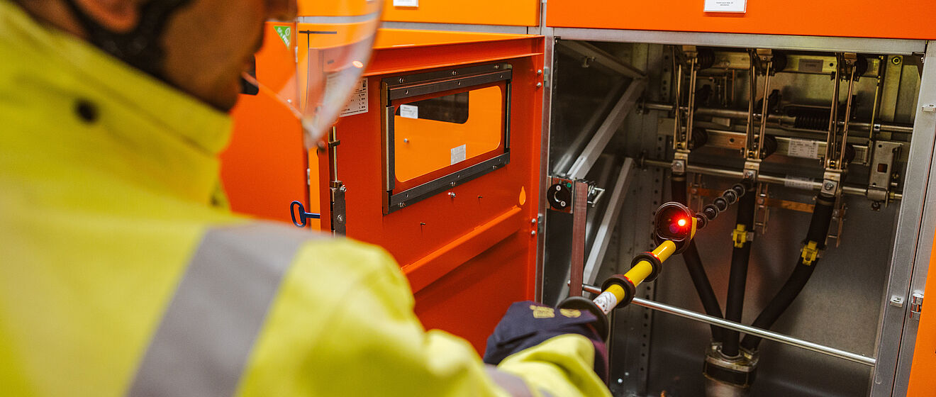 Elektrofachkraft führt Schalthandlung an einer Mittelspannungsanlage durch: Die Person trägt Warnschutzkleidung und einen Schutzhelm und bedient mit einer isolierten Schaltstange eine geöffnete orangefarbene Schaltanlage. Im Hintergrund sind weitere geschlossene Schaltschränke sichtbar. Die Szene zeigt sicheres Arbeiten an elektrischen Anlagen im Innenbereich. Verlinkung zur Kabelfehlerortung Seite.