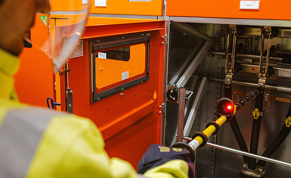 Elektrofachkraft führt Schalthandlung an einer Mittelspannungsanlage durch: Die Person trägt Warnschutzkleidung und einen Schutzhelm und bedient mit einer isolierten Schaltstange eine geöffnete orangefarbene Schaltanlage. Im Hintergrund sind weitere geschlossene Schaltschränke sichtbar. Die Szene zeigt sicheres Arbeiten an elektrischen Anlagen im Innenbereich. Verlinkung zur Kabelfehlerortung Seite.