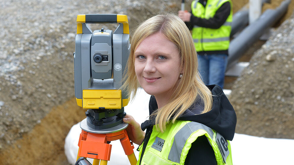 Auf einer Baustelle steht eine Person in einer gelben Warnweste vor einem Vermessungsgerät auf einem Stativ. Die Person hält ein Steuergerät in der Hand und bedient das Messinstrument. Im Hintergrund ist eine weitere Person in Warnweste zu sehen, die eine Messlatte hält. Der Boden besteht aus Kies und Erde, und im Graben liegen Rohre. Die Szene zeigt Vermessungsarbeiten für Bau- oder Infrastrukturprojekte.