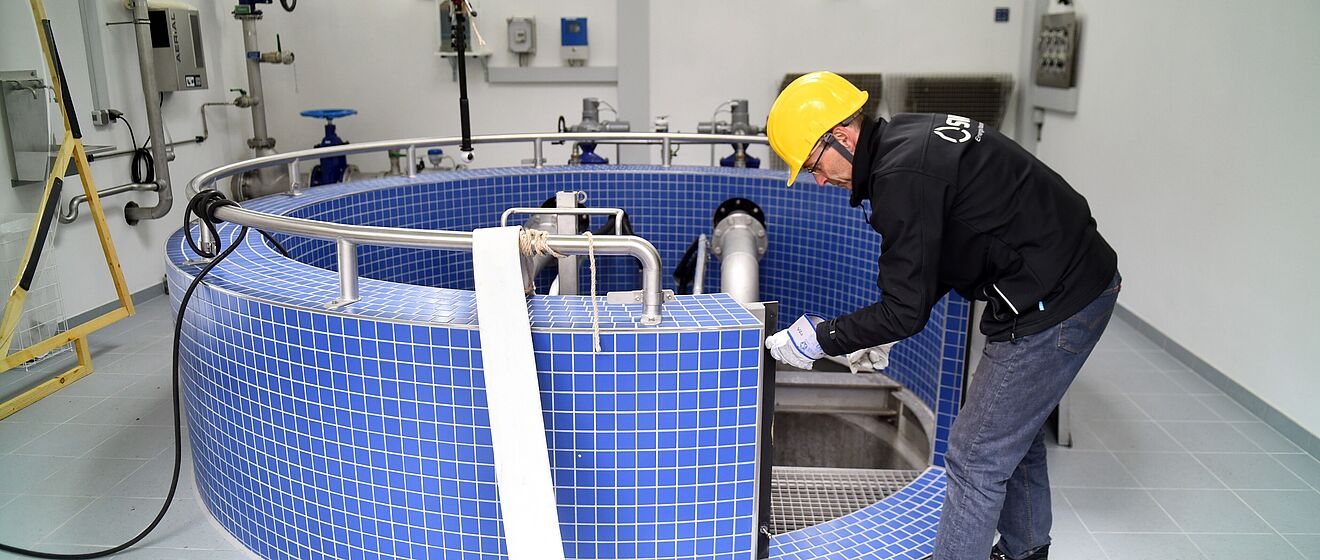 Eine Person trägt einen gelben Schutzhelm und schwarze Arbeitskleidung und arbeitet in einem Technikraum an einer großen runden Anlage mit blauen Fliesen. Die Anlage ist mit Edelstahlrohren und Armaturen ausgestattet, die in den Innenbereich führen. Im Hintergrund sind weiße Wände mit montierten Steuerkästen, Rohrleitungen und Ventilen sichtbar. Die Beleuchtung ist hell und gleichmäßig. Verlinkung zur Wasser Seite.