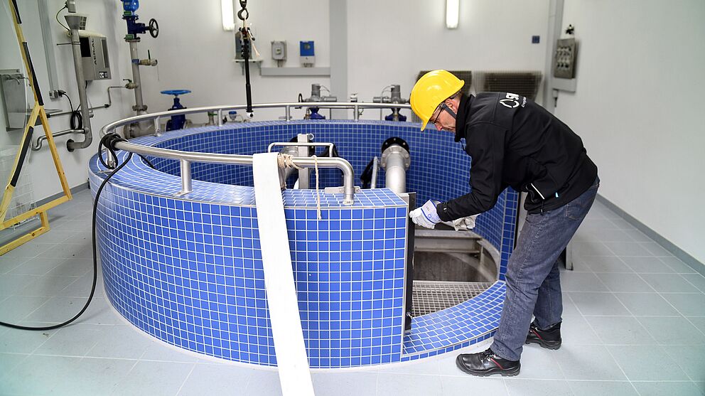 Eine Person trägt einen gelben Schutzhelm und schwarze Arbeitskleidung und arbeitet in einem Technikraum an einer großen runden Anlage mit blauen Fliesen. Die Anlage ist mit Edelstahlrohren und Armaturen ausgestattet, die in den Innenbereich führen. Im Hintergrund sind weiße Wände mit montierten Steuerkästen, Rohrleitungen und Ventilen sichtbar. Die Beleuchtung ist hell und gleichmäßig. Verlinkung zur Wasser Seite.