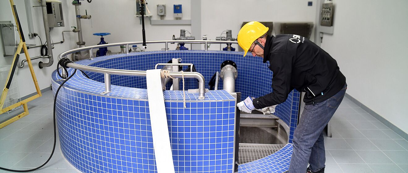 Eine Person trägt einen gelben Schutzhelm und schwarze Arbeitskleidung und arbeitet in einem Technikraum an einer großen runden Anlage mit blauen Fliesen. Die Anlage ist mit Edelstahlrohren und Armaturen ausgestattet, die in den Innenbereich führen. Im Hintergrund sind weiße Wände mit montierten Steuerkästen, Rohrleitungen und Ventilen sichtbar. Die Beleuchtung ist hell und gleichmäßig. Verlinkung zur Wasser Seite.