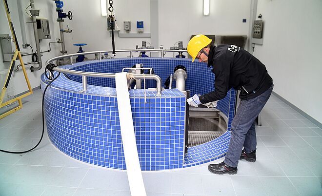 Eine Person trägt einen gelben Schutzhelm und schwarze Arbeitskleidung und arbeitet in einem Technikraum an einer großen runden Anlage mit blauen Fliesen. Die Anlage ist mit Edelstahlrohren und Armaturen ausgestattet, die in den Innenbereich führen. Im Hintergrund sind weiße Wände mit montierten Steuerkästen, Rohrleitungen und Ventilen sichtbar. Die Beleuchtung ist hell und gleichmäßig. Verlinkung zur Wasser Seite.