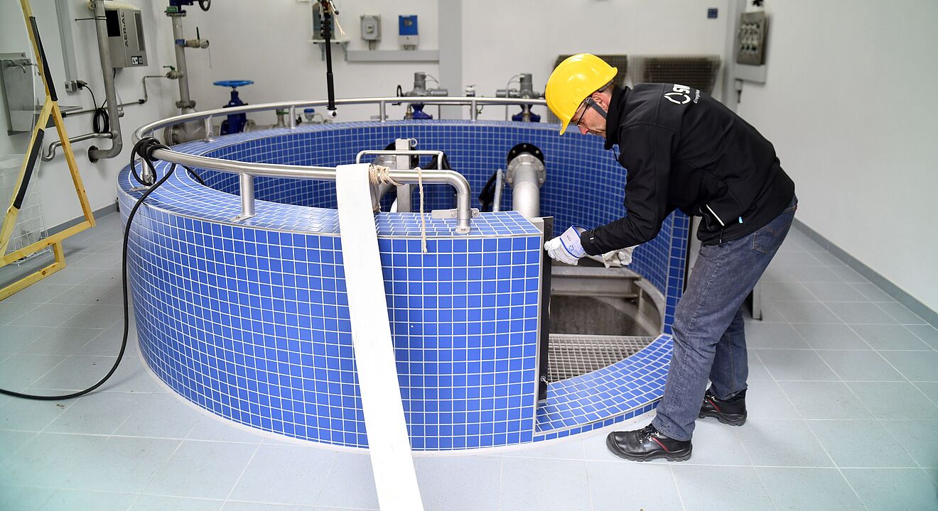 Eine Person trägt einen gelben Schutzhelm und schwarze Arbeitskleidung und arbeitet in einem Technikraum an einer großen runden Anlage mit blauen Fliesen. Die Anlage ist mit Edelstahlrohren und Armaturen ausgestattet, die in den Innenbereich führen. Im Hintergrund sind weiße Wände mit montierten Steuerkästen, Rohrleitungen und Ventilen sichtbar. Die Beleuchtung ist hell und gleichmäßig. Verlinkung zur Wasser Seite.