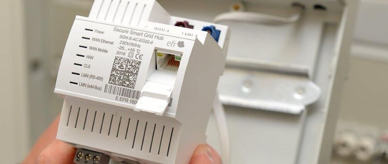 Eine Hand hält ein weißes Smart-Grid-Modul mit der Aufschrift ‚Secure Smart Grid Hub‘ und sichtbarem QR-Code. Das Gerät verfügt über mehrere Anschlüsse für WAN, Ethernet und LAN. Im Hintergrund ist ein Stromzähler mit dem Logo der swa Netze montiert. Die Szene zeigt moderne Messtechnik für intelligente Stromnetze. Verlinkung zur Smart Meter Seite.