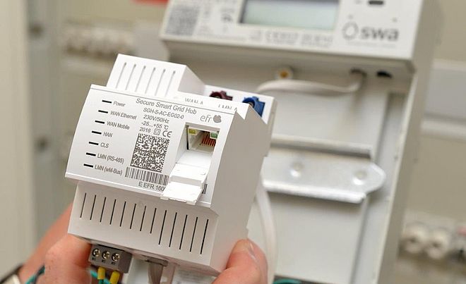 Eine Hand hält ein weißes Smart-Grid-Modul mit der Aufschrift ‚Secure Smart Grid Hub‘ und sichtbarem QR-Code. Das Gerät verfügt über mehrere Anschlüsse für WAN, Ethernet und LAN. Im Hintergrund ist ein Stromzähler mit dem Logo der swa Netze montiert. Die Szene zeigt moderne Messtechnik für intelligente Stromnetze. Verlinkung zur Smart Meter Seite.