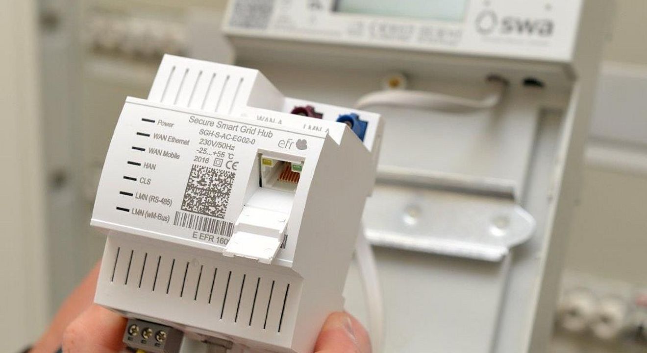 Eine Hand hält ein weißes Smart-Grid-Modul mit der Aufschrift ‚Secure Smart Grid Hub‘ und sichtbarem QR-Code. Das Gerät verfügt über mehrere Anschlüsse für WAN, Ethernet und LAN. Im Hintergrund ist ein Stromzähler mit dem Logo der swa Netze montiert. Die Szene zeigt moderne Messtechnik für intelligente Stromnetze. Verlinkung zur Smart Meter Seite.