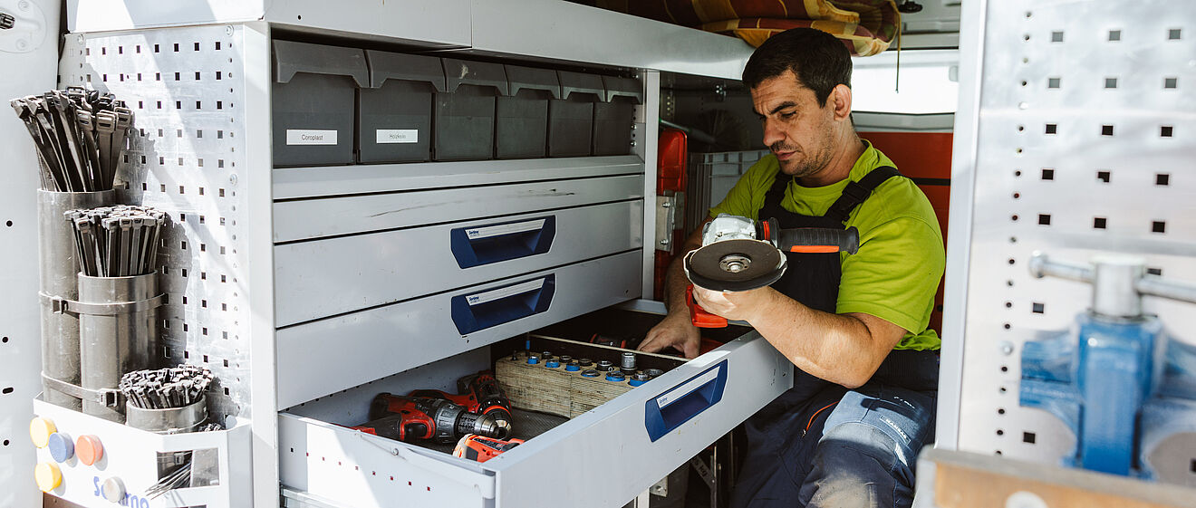 Innenansicht eines Servicefahrzeugs mit einer professionellen Werkstatteinrichtung. Links sind Regale mit Sortierboxen, Kabelbindern und Werkzeugen zu sehen. In der Mitte befindet sich eine geöffnete Schublade mit Akkuschraubern, Werkzeugen und Kleinteilen. Eine Person kniet vor der Schublade und hält ein rundes Ersatzteil in der Hand. Die Einrichtung ist sauber und gut organisiert, mit dem Markenlogo ‚Sortimo‘ auf der rechten Seite. Verlinkung zur Hausanschluss Seite.