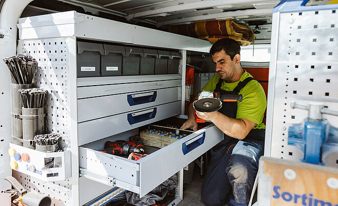 Innenansicht eines Servicefahrzeugs mit einer professionellen Werkstatteinrichtung. Links sind Regale mit Sortierboxen, Kabelbindern und Werkzeugen zu sehen. In der Mitte befindet sich eine geöffnete Schublade mit Akkuschraubern, Werkzeugen und Kleinteilen. Eine Person kniet vor der Schublade und hält ein rundes Ersatzteil in der Hand. Die Einrichtung ist sauber und gut organisiert, mit dem Markenlogo ‚Sortimo‘ auf der rechten Seite. Verlinkung zur Hausanschluss Seite.