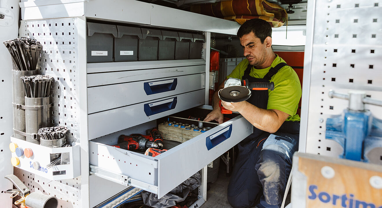 Innenansicht eines Servicefahrzeugs mit einer professionellen Werkstatteinrichtung. Links sind Regale mit Sortierboxen, Kabelbindern und Werkzeugen zu sehen. In der Mitte befindet sich eine geöffnete Schublade mit Akkuschraubern, Werkzeugen und Kleinteilen. Eine Person kniet vor der Schublade und hält ein rundes Ersatzteil in der Hand. Die Einrichtung ist sauber und gut organisiert, mit dem Markenlogo ‚Sortimo‘ auf der rechten Seite. Verlinkung zur Hausanschluss Seite.