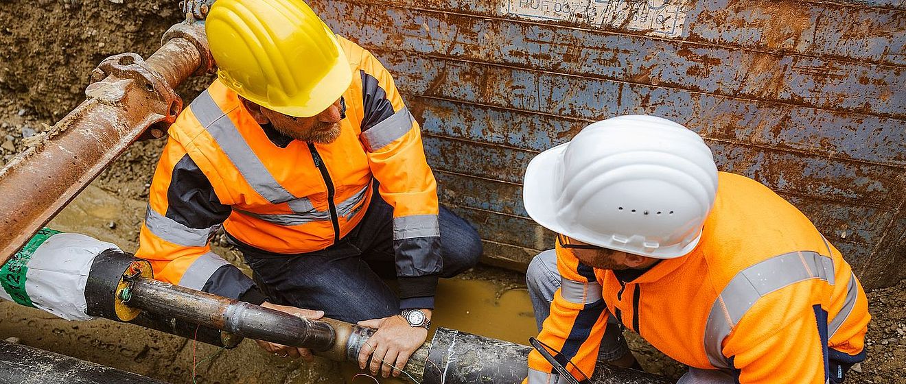Zwei Fachkräfte in hochsichtbarer orangefarbener Schutzkleidung mit reflektierenden Streifen und Schutzhelmen arbeiten in einer Baugrube an unterirdischen Versorgungsleitungen. Im Vordergrund verlaufen mehrere schwarze Rohrleitungen parallel durch feuchten, schlammigen Boden. Eine Person stabilisiert eine Rohrverbindung, während die andere Person mit einem Werkzeug eine Verbindung oder Messung überprüft. Die Rohrabschnitte zeigen Muffen und Übergangsstücke, teilweise mit farbigen Markierungen und freiliegenden Leitungen. Im Hintergrund steht ein großer, rostiger Metallcontainer oder Baukasten mit Kratzspuren. Die Szene vermittelt Wartungs- und Reparaturarbeiten an kritischer Infrastruktur wie Wasser-, Gas- oder Fernwärmenetzen unter realen Baustellenbedingungen. Verlinkung zur swa Netze Baustellen Seite.