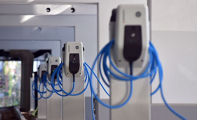 Reihe moderner Ladestationen für Elektrofahrzeuge in einem überdachten Bereich: Die weißen Wallboxen sind auf Metallpfosten montiert und mit blauen Ladekabeln ausgestattet, die ordentlich aufgewickelt sind. Der Boden ist mit Laub bedeckt, im Hintergrund sind weitere Ladestationen sichtbar. Die Szene symbolisiert Ladeinfrastruktur für Elektromobilität. Verlinkung zur emobilitaet Seite