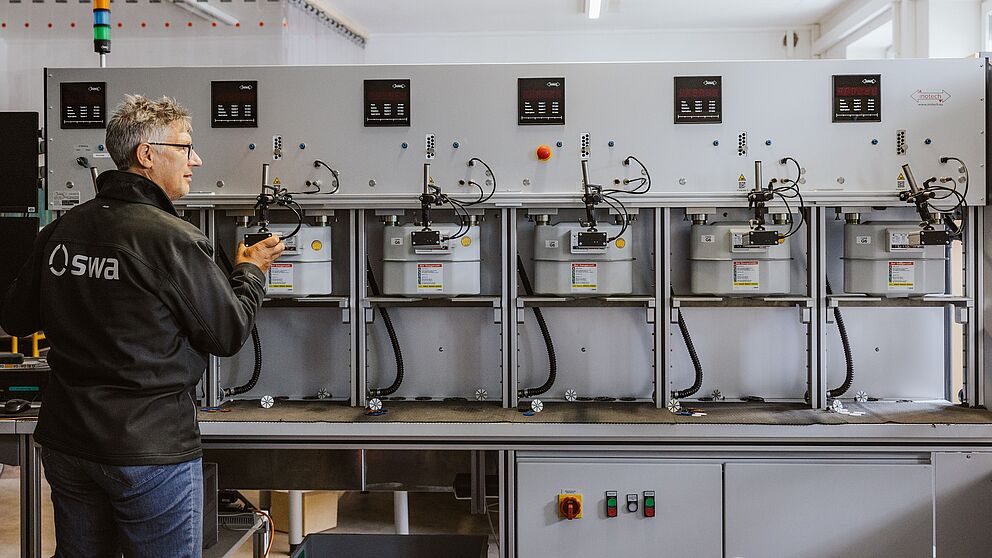 Prüfanlage für Gaszähler mit sechs montierten Gaszählern in einer Reihe. Die Anlage besteht aus einer grauen Metallkonstruktion mit Anzeigen, Bedienelementen und Anschlüssen für Messleitungen. Links steht eine Person in Arbeitskleidung mit dem Logo ‚swa‘ auf der Rückseite und bedient die Anlage.