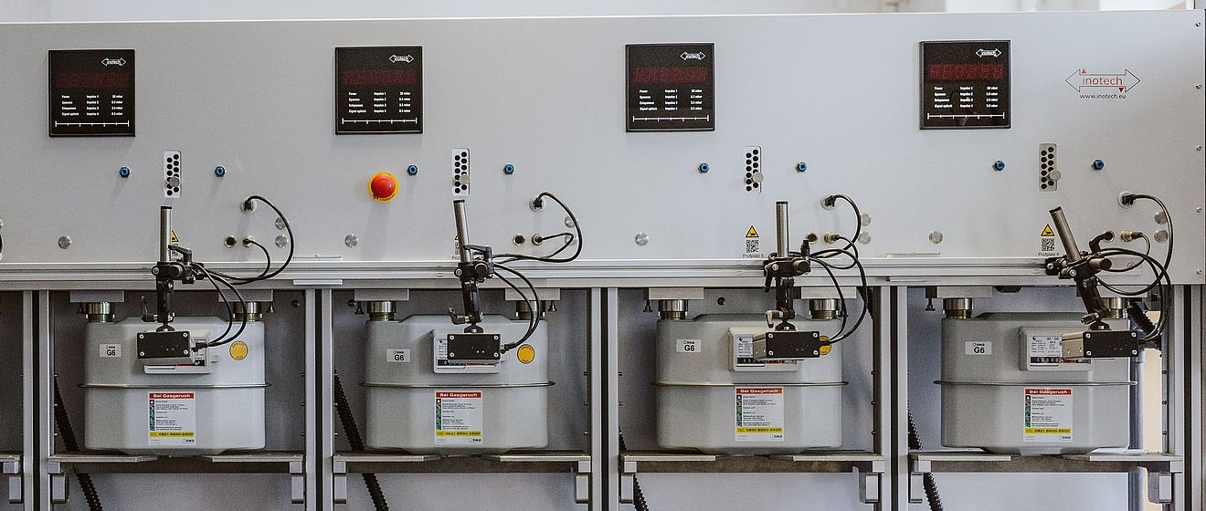 Prüfanlage für Gaszähler mit sechs montierten Gaszählern in einer Reihe. Die Anlage besteht aus einer grauen Metallkonstruktion mit Anzeigen, Bedienelementen und Anschlüssen für Messleitungen. Links steht eine Person in Arbeitskleidung mit dem Logo ‚swa‘ auf der Rückseite und bedient die Anlage.