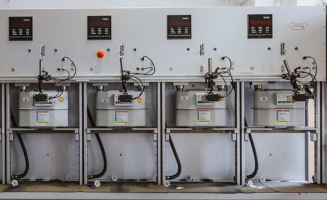 Prüfanlage für Gaszähler mit sechs montierten Gaszählern in einer Reihe. Die Anlage besteht aus einer grauen Metallkonstruktion mit Anzeigen, Bedienelementen und Anschlüssen für Messleitungen. Links steht eine Person in Arbeitskleidung mit dem Logo ‚swa‘ auf der Rückseite und bedient die Anlage.