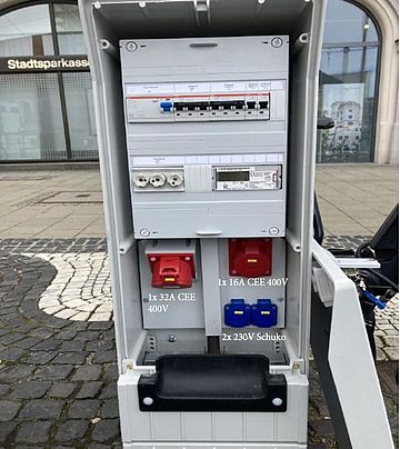 Geöffneter mobiler Stromverteiler der swa Netze GmbH im öffentlichen Raum. Im Schaltschrank sind Sicherungen, Fehlerstromschutzschalter und ein digitaler Stromzähler integriert. Im unteren Bereich befinden sich ein 32A-CEE-400V-Anschluss, ein 16A-CEE-400V-Anschluss sowie zwei 230V-Schuko-Steckdosen. Die robuste Verteilereinheit ermöglicht eine sichere und flexible Stromversorgung für Veranstaltungen, Baustellen oder temporäre Einsatzzwecke. Im Hintergrund sind Pflasterflächen und ein Gebäude zu sehen.