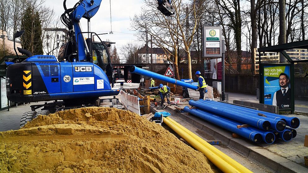 Eine Baustelle im städtischen Bereich mit einem großen blauen Bagger, der ein langes blaues Rohr hält. Auf dem Boden liegen mehrere blaue und gelbe Rohre nebeneinander. Im Vordergrund befindet sich ein großer Sandhaufen. Mehrere Personen in Warnwesten arbeiten an der Baustelle, die mit rot-weißen Absperrungen gesichert ist. Im Hintergrund sind eine Bushaltestelle mit einem Fahrplan, ein Werbeplakat und Bäume zu sehen. Die Szene zeigt Rohrleitungsarbeiten für Infrastruktur oder Versorgungsnetze.