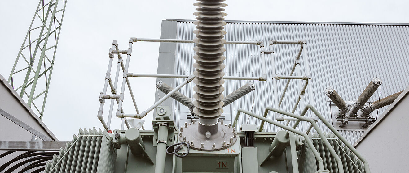 Großer Hochspannungstransformator in einem Umspannwerk: Im Vordergrund ist die Oberseite des Transformators mit einer zentralen Isolator-Durchführung und mehreren Anschlussarmen sichtbar. Die Anlage ist grau lackiert und mit metallischen Leitungen und Isolatoren ausgestattet. Im Hintergrund sind Gebäudewände, ein Stahlgittermast und weitere technische Komponenten erkennbar. Die Szene zeigt moderne Energieinfrastruktur für Stromübertragung und Netztechnik.