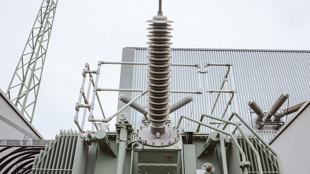 Großer Hochspannungstransformator in einem Umspannwerk: Im Vordergrund ist die Oberseite des Transformators mit einer zentralen Isolator-Durchführung und mehreren Anschlussarmen sichtbar. Die Anlage ist grau lackiert und mit metallischen Leitungen und Isolatoren ausgestattet. Im Hintergrund sind Gebäudewände, ein Stahlgittermast und weitere technische Komponenten erkennbar. Die Szene zeigt moderne Energieinfrastruktur für Stromübertragung und Netztechnik.