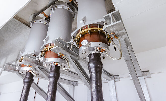 Nahaufnahme von drei Hochspannungs-Kabelendverschlüssen in einer elektrischen Anlage. Die Kabel sind schwarz isoliert und führen in massive metallische Endverschlüsse, die mit Kupfer- und Messingelementen sowie Schraubverbindungen fixiert sind. Die Konstruktion ist auf stabilen Metallträgern montiert und dient der sicheren Verbindung und Isolation von Hochspannungskabeln. Die Umgebung ist sauber und technisch ausgeführt, typisch für einen geschlossenen Schaltraum. Verlinkung zur Einspeisung Seite.