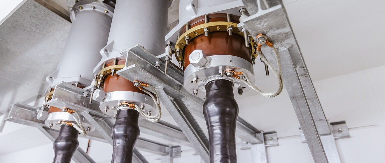 Nahaufnahme von drei Hochspannungs-Kabelendverschlüssen in einer elektrischen Anlage. Die Kabel sind schwarz isoliert und führen in massive metallische Endverschlüsse, die mit Kupfer- und Messingelementen sowie Schraubverbindungen fixiert sind. Die Konstruktion ist auf stabilen Metallträgern montiert und dient der sicheren Verbindung und Isolation von Hochspannungskabeln. Die Umgebung ist sauber und technisch ausgeführt, typisch für einen geschlossenen Schaltraum. Verlinkung zur Einspeisung Seite.