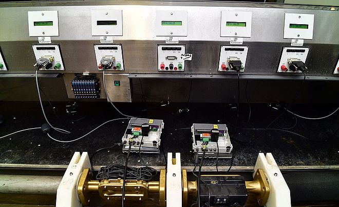 Prüfstand für elektronische Wärmezähler in einem Labor der Versorgungsinfrastruktur. Im Vordergrund sind zwei geöffnete Wärmezähler auf Halterungen montiert und über Kabel mit Mess- und Prüfgeräten verbunden. Im Hintergrund befindet sich eine Edelstahl-Prüfwand mit mehreren digitalen Anzeigen, Anschlussfeldern und Messschnittstellen zur parallelen Kalibrierung und Funktionsprüfung. Die technische Anlage dient der Qualitätskontrolle, Parametrierung und Verifizierung von Messgeräten im Netzbetrieb.