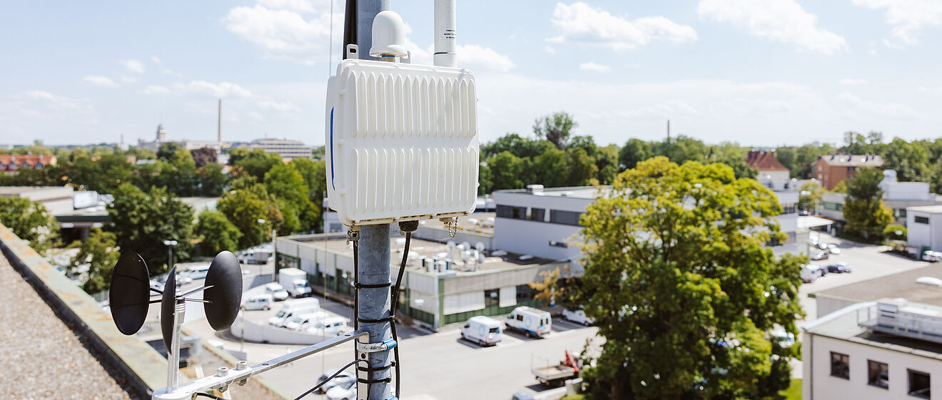 Auf einem Dach ist ein technisches Mess- und Kommunikationsgerät an einem Mast befestigt. Das weiße Gehäuse enthält Antennen und Kabel, darunter ist ein Windmessgerät mit zwei Schalen und einem Windrichtungsanzeiger montiert. Im Hintergrund sind Gebäude, Bäume und parkende Fahrzeuge zu sehen, bei klarem Himmel mit vereinzelten Wolken. Verlinkung zur IoT Seite.